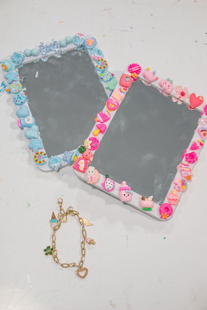Two rectangular mirrors bordered with colorful, playful clay decorations, including ice creams, fruits, and hearts. One frame is blue-themed, the other pink. Below the mirrors lies a gold bracelet with various charms. They rest on a white surface.